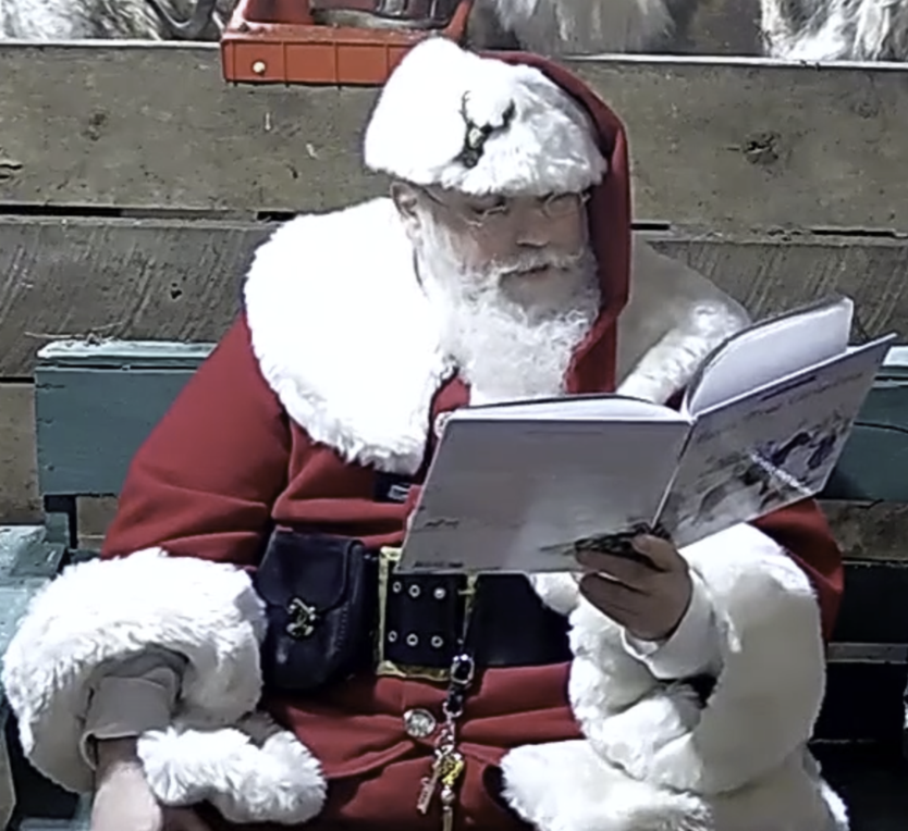 Santa sitting in green chair looking at book he is holding in his left hand